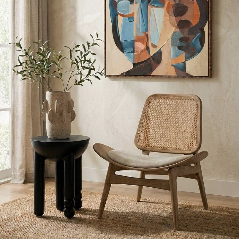 An abstract framed canvas print with geometric shapes in blue, beige, brown, and black, displayed on a textured beige wall in a styled room setting with a wooden chair, a black side table, and a potted plant.