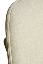 Close-up of a light beige textured fabric cushion with a curved edge and a small portion of a brown frame or backing visible on the left side, set against a plain white background.