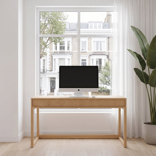 A light natural wood writing desk with a single drawer and slatted front, styled in a bright home office with a computer monitor on top, a large window showing buildings outside, and a potted plant to the right.