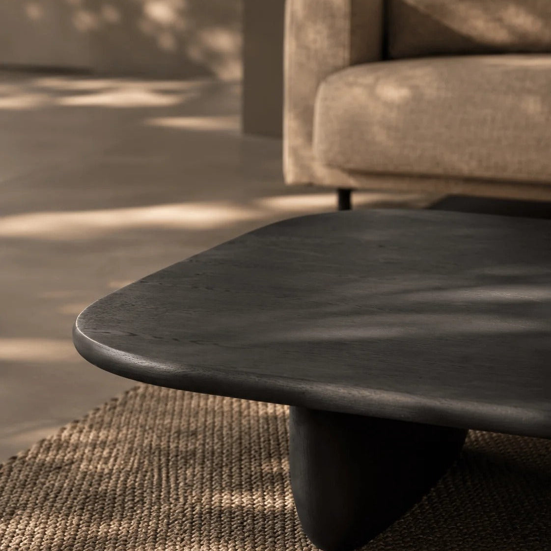 A dark charcoal-colored wooden coffee table with a rounded organic edge and a thick sculptural base, set on a textured beige rug in a modern living room with dappled sunlight.