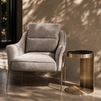 A modern gray upholstered armchair with slim gold-toned legs sits beside a cylindrical side table with a brushed brass base and black top, set in a sunlit room with dappled shadows on a textured beige wall and floor.