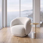 A modern round armchair upholstered in textured cream bouclé fabric, positioned in a bright room with a wooden floor and a small round gold side table next to it. Large windows behind the chair offer a scenic view of a misty coastal ocean.